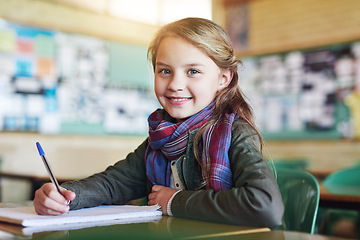 Image showing Education, portrait of young girl in classroom and writing in her notebook at her desk in school building. Studying or homework, learn or academic and happy female student with pen sitting on chair