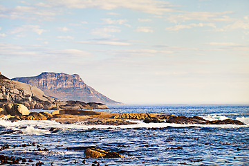 Image showing Mountains, ocean and nature with waves at beach for environment, landscape and blue sky. Calm, summer and seascape with sunrise on horizon at coastline for tropical, clouds and water on rock surface