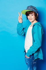 Image showing Fashion, peace sign and portrait of a boy in a studio with a casual, trendy and cool outfit. Teenager, smile and happy boy kid model with hipster apparel style posing isolated by a blue background.