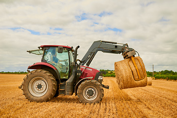 Image showing Hay, agriculture and a red tractor on a farm for sustainability outdoor on an open field during the harvest season. Nature, sky and clouds with an agricultural vehicle harvesting in the countryside