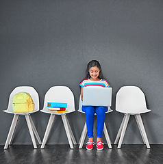 Image showing Child, chair and laptop for learning and education while typing for research on internet. Happy girl kid or student against a grey wall with technology, book and backpack in a waiting room with space