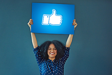 Image showing Portrait, social media and thumbs up icon to like with a woman holding a poster in studio on a blue background. Smile, logo and button with a happy female influencer showing a hand gesture sign