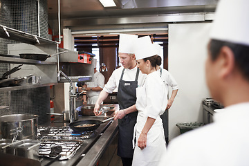 Image showing Chef, teamwork and people with frying pan in kitchen, catering service and prepare food for fine dining. Professional, cooking and man training woman, saute and cook add oil to meat in restaurant.