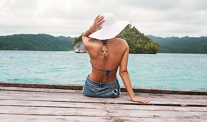 Image showing Travel, hat and back of woman on an island to relax while on tropical summer vacation or holiday. Adventure, outdoor and female person sitting on wood pier by ocean water on weekend trip in paradise.