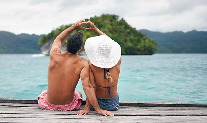 Image showing Heart hands, back and couple by a ocean while on summer, paradise and tropical vacation. Romance, dock and young man and woman relaxing on wood pier with love shape gesture on holiday or weekend trip