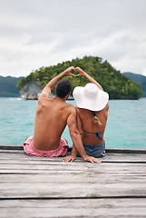 Image showing Heart shape, hands and couple by ocean while on a summer, romantic and tropical vacation. Island, dock and back of man and woman relaxing on wood pier with love gesture on holiday or weekend trip.