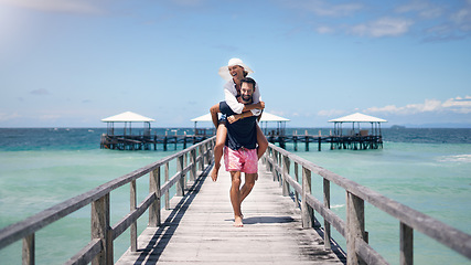 Image showing Ocean, dock and a couple playing on vacation for a piggy back, freedom and to relax outdoor with love. A man and woman together on holiday, tropical travel and summer adventure in nature with water