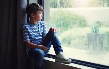 Image showing Little boy, home and watching the rain by window of natural water drops and thinking in relax indoors. Child relaxing by windowsill looking out glass at storm in lonely or sad for weather at house