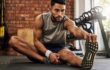 Image showing Gym, man sitting and stretching legs for workout warm up, motivation and fitness mindset with hand on foot. Focus, commitment and male athlete on floor, leg stretch at sports club for exercise goals