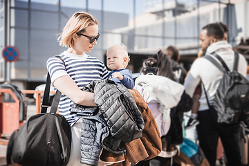 Image showing Motherat travelling with his infant baby boy child. Mom holding travel bag and her infant baby boy child while queuing for bus in front of airport terminal station.