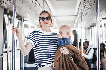 Image showing Mother carries her child while standing and holding on to bar holder on bus. Mom holding infant baby boy in her arms while riding in public transportation. Cute toddler traveling with mother.