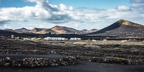 Image showing Traditional white houses in black volcanic landscape of La Geria wine growing region with view of Timanfaya National Park in Lanzarote. Touristic attraction in Lanzarote island, Canary Islands, Spain.