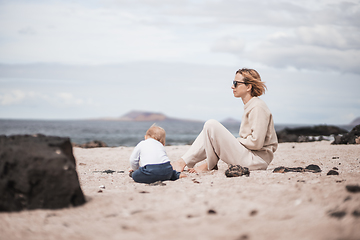 Image showing Mother enjoying winter beach vacations playing with his infant baby boy son on wild volcanic sandy beach on Lanzarote island, Canary Islands, Spain. Family travel and vacations concept