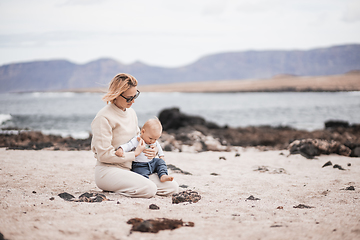 Image showing Mother enjoying winter beach vacations playing with his infant baby boy son on wild volcanic sandy beach on Lanzarote island, Canary Islands, Spain. Family travel and vacations concept