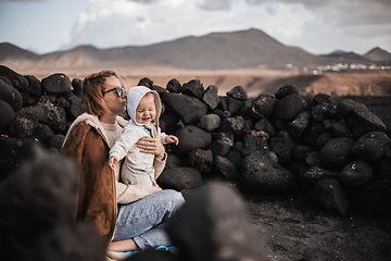 Image showing Mother enjoying winter vacations playing with his infant baby boy son on black sandy volcanic beach of Janubio on Lanzarote island, Spain on windy overcast day. Family travel vacations concept.