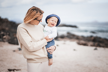 Image showing Mother enjoying winter vacations holding and playing with his infant baby boy son on volcanic sandy beach on Lanzarote island, Spain. Family travel and vacations concept