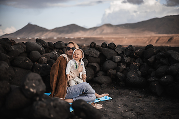 Image showing Mother enjoying winter vacations playing with his infant baby boy son on black sandy volcanic beach of Janubio on Lanzarote island, Spain on windy overcast day. Family travel vacations concept.