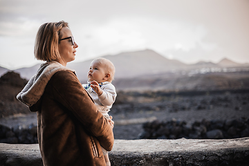 Image showing Mother enjoying winter vacations playing with his infant baby boy son on black sandy volcanic beach of Janubio on Lanzarote island, Spain on windy overcast day. Family travel vacations concept.