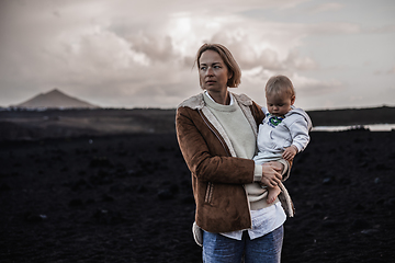 Image showing Mother enjoying winter vacations playing with his infant baby boy son on black sandy volcanic beach of Janubio on Lanzarote island, Spain on windy overcast day. Family travel vacations concept.