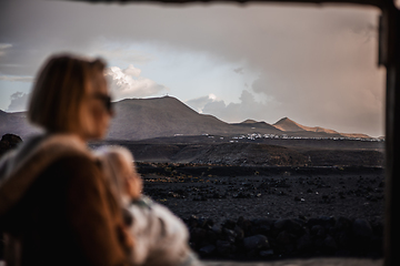 Image showing Mother enjoying winter vacations playing with his infant baby boy son on black sandy volcanic beach of Janubio on Lanzarote island, Spain on windy overcast day. Focus on landscape in background.