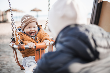 Image showing Mother pushing her cheerful infant baby boy child on a swing on sandy beach playground outdoors on nice sunny cold winter day in Malaga, Spain.