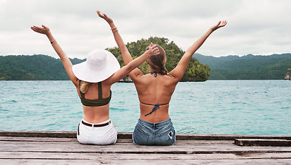 Image showing Rear view, people sitting on a jetty and carefree for excitement outdoors by the ocean. Summer vacation or holiday break, freedom and excited or cheerful women sit on pier at sea for fresh air