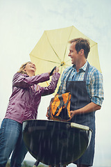 Image showing Man, woman and outdoor for bbq in rain with umbrella, charcoal and surprise on vacation in low angle. Couple, outdoor and grill in storm, cooking and insurance in winter with comic laughing in garden