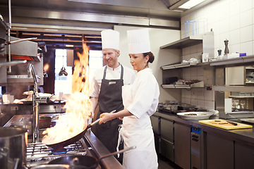 Image showing Chef, people and fire with frying pan in kitchen, catering service and prepare food for fine dining. Professional, cooking and man and woman flambe and smile for teamwork in commercial restaurant