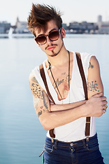 Image showing Sunglasses, portrait and man by sea with arms crossed for stylish tattoo, body art and fashion. Hipster, ocean and confident male person standing outdoor in Spain with cool clothes, attitude and punk