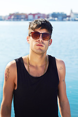 Image showing Portrait, sunglasses and serious man by ocean with stylish tattoo, body art and fashion. Face, sea and trendy male person standing outdoor in Norway with cool clothes, attitude and youth aesthetic.