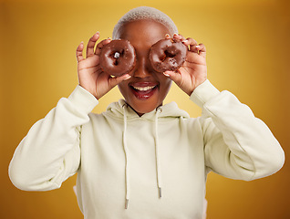 Image showing Face, chocolate and donut with a black woman in studio on a gold background for candy or unhealthy eating. Smile, food and baking with an excited young female person holding sweet pastry for dessert