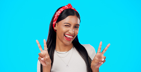 Image showing Face, funny and Indian woman with peace sign, confident and happiness on a blue studio background. Portrait, female and lady with smile, silly and goofy with wink, joyful and cheerful with excitement