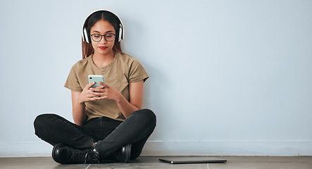 Image showing Music headphones, floor and woman with phone in home by wall background with mockup. Cellphone, social media and female sitting on ground while streaming or listening to podcast, radio sound or audio