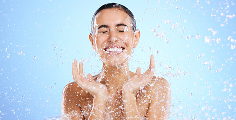 Image showing Water splash, face and woman shower in studio, blue background and healthy beauty of wellness skincare. Happy female model, wet drops and cleaning of facial, aqua hydration or smile on color backdrop