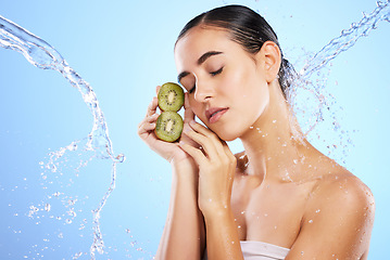 Image showing Woman, water splash and face with kiwi in relax for skincare, hygiene or nutrition against blue studio background. Calm or relaxed female holding fruit in natural beauty cosmetics or facial treatment