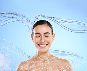 Image showing Water splash, smile and woman cleaning in studio, blue background and healthy beauty of wellness skincare. Happy female model, wet and shower for hygiene, facial hydration and face on color backdrop