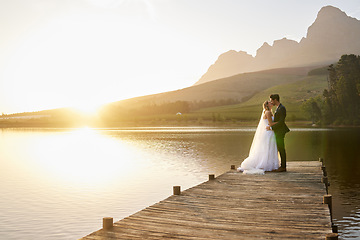 Image showing Kiss, sunset and married couple by a lake for a wedding, marriage ceremony and event. Together, happy and a bride and groom standing by a pier, kissing and giving affection to celebrate their love