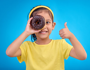 Image showing Donut, eye and thumbs up with portrait of girl in studio for junk food, sugar and happiness. Snack, cake and cute with face of child and dessert for eating, positive and chocolate on blue background