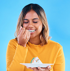 Image showing Birthday, blue background and a woman eating cake in studio while having fun at a party for celebration. Dessert, food and fork with an attractive young female enjoying a rainbow pastry snack