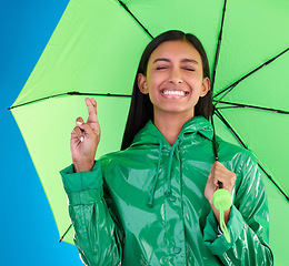 Image showing Green, umbrella and fingers crossed with a woman in studio on a blue background during winter for insurance. Rain, fashion or cover and an attractive young female wishing for weather change with hope