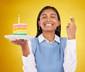 Image showing Smile, wish and woman with cake in studio for happy celebration or party on yellow background. Happiness, excited gen z model with fingers crossed and candle in rainbow dessert to celebrate milestone