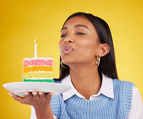 Image showing Birthday, cake and woman blowing candle in studio for happy party celebration on yellow background. Happiness, gen z girl and candles in rainbow dessert to celebrate milestone, event or achievement.