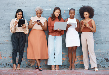 Image showing Technology, diversity and business women by a wall standing in a line doing research online. Professional females with phone, tablet and laptop working on a resume while waiting for a job interview.