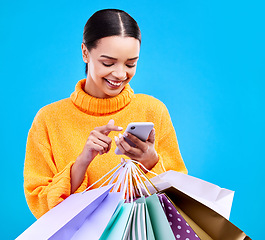 Image showing Happy woman, phone and shopping bags with smile for purchase, sale or discount against a blue studio background. Excited female shopper smiling on mobile smartphone holding gift bag for social media