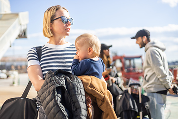 Image showing Motherat travelling with his infant baby boy child. Mom holding travel bag and her infant baby boy child while queuing for bus in front of airport terminal station.