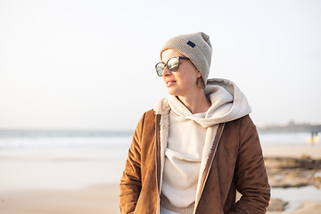 Image showing Portrait of young stylish woman wearing brown padded jacket, hoodie, wool cap and sunglasses on long sandy beach in spring.