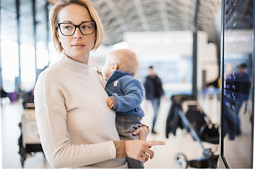 Image showing Mother traveling with child, holding his infant baby boy at airport terminal, checking flight schedule, waiting to board a plane. Travel with kids concept.