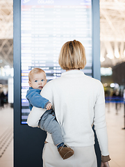 Image showing Mother traveling with child, holding his infant baby boy at airport terminal, checking flight schedule, waiting to board a plane. Travel with kids concept.