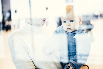 Image showing Thoughtful young mother looking trough window holding his infant baby boy child while waiting to board an airplane at airport terminal departure gates. Travel with baby concept.