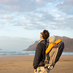 Image showing Young father rising hands to the sky while enjoying pure nature carrying his infant baby boy son in backpack on windy sandy beach of Famara, Lanzarote island, Spain at sunset. Family travel concept.